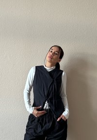 Giovane donna con capelli scuri raccolti, che indossa un gilet senza maniche nero sopra una camicia bianca a maniche lunghe e pantaloni neri larghi, posata contro un muro neutro.