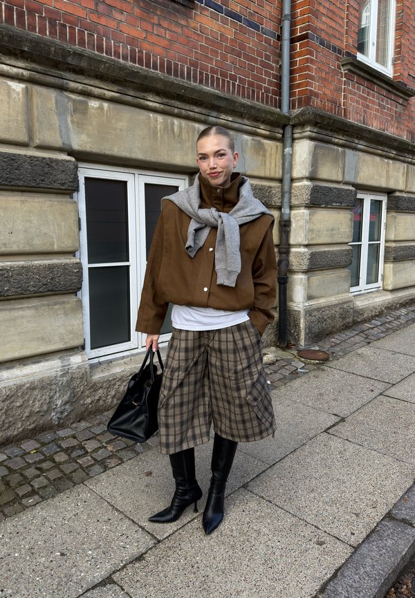 Žena stojaca na chodníku v hnedej bunde, károvaných širokých nohaviciach, čiernych čižmách, sivom svetri prehodenom cez ramená, drží čiernu kabelku.