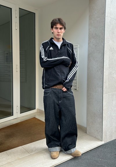 Chaqueta negra con cremallera y rayas blancas, camisa blanca, jeans negros holgados y zapatos marrones claros. De pie en una puerta con azulejos grises y vidrio.