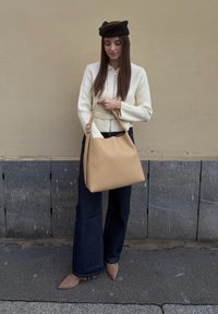 Femme portant un béret noir, un cardigan crème à fermeture éclair, un jean large foncé et des chaussures beige pointues, tenant un grand sac à main marron clair.