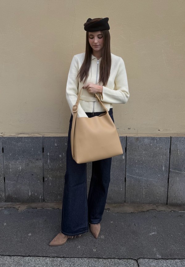 Femme portant un béret noir, un cardigan crème à fermeture éclair, un jean large foncé et des chaussures beiges pointues, tenant un grand sac à main marron clair.