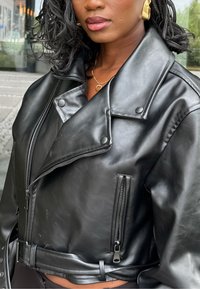 Musta keinonahkainen lyhyt takki, jossa on iso kaulus, vetoketjulliset taskut ja nepparit; vyötäisillä vyö ja sileä pinta.