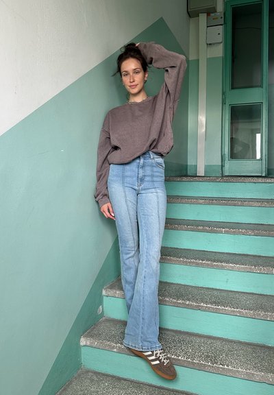 Suéter marrón de gran tamaño, jeans acampanados de tiro alto en azul claro y zapatillas marrones con rayas blancas, de pie en escaleras verdes y grises.