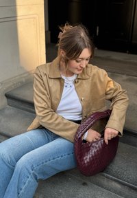 Vevet bordeaux håndveske med en buet håndtak, kombinert med en beige semsket skinnjakke og blå jeans, sittende på steintrapper.