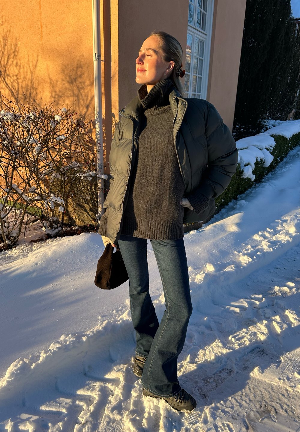 Puffermantel in dunklem Grün, Rollkragenpullover in Anthrazitgrau, ausgestellte Jeans in Blau, schwarze Tasche und schwarze Schuhe, vor schneebedecktem Boden.
