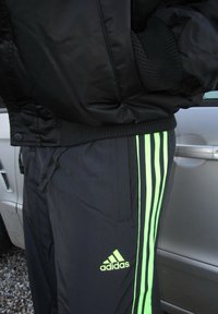 Blusão bomber preto usado com calças pretas que apresentam três riscas verticais em verde neón e um logótipo da Adidas na perna esquerda.