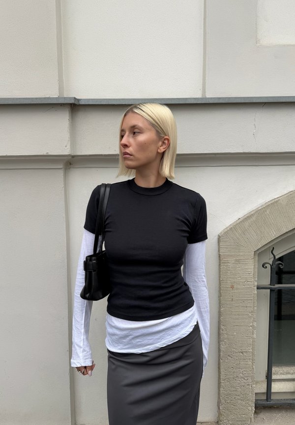 Mujer con cabello rubio corto que lleva puestos tops en capas de color negro y blanco y una falda gris, llevando un bolso negro de hombro, de pie frente a una pared beige.