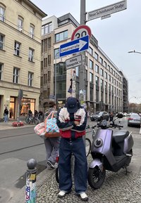 Homme portant une casquette et une veste, appuyé sur un poteau avec des panneaux de signalisation, un scooter à ses côtés, et des piétons marchant dans la rue de la ville devant des bâtiments.