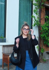 Czarna marynarka skrojona na miarę noszona na czarnej bluzce, w zestawieniu z dżinsową spódnicą. Dodatki to czarna torebka i srebrny zegarek, w tle ogrodowa sceneria.