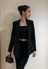 Femme en blazer noir et crop top tenant un petit sac à main noir, regardant vers la droite sur un fond clair uni.