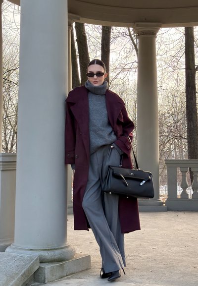 Mujer con gafas de sol que lleva un abrigo burdeos, un suéter de cuello alto gris y pantalones grises, apoyada contra una columna en un pabellón al aire libre.