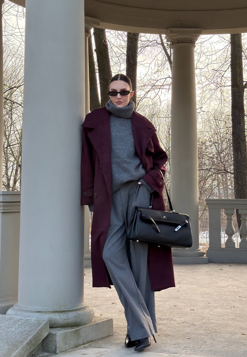 Donna con occhiali da sole che indossa un cappotto color borgogna, dolcevita grigio e pantaloni grigi, appoggiata a una colonna in un padiglione all'aperto.