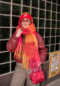 Piros bőr dzseki, többszínű gyapjúsál rojttal, barna nadrág, kis piros kézitáska, kerek napszemüveg és piros baseballsapka.