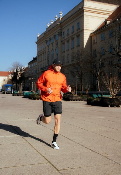 Zapatillas de correr, pantalones cortos negros, chaqueta naranja y gorra negra. La chaqueta tiene un cierre frontal; los pantalones cortos tienen un logo. Luz solar brillante, fondo urbano.
