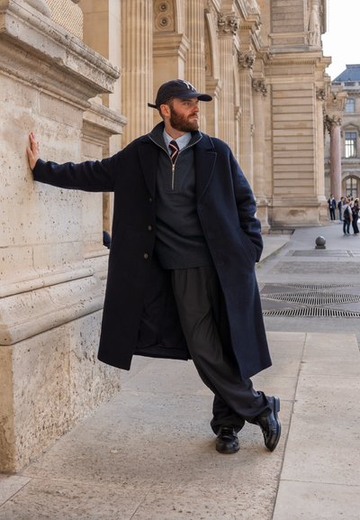 Hombre vistiendo un abrigo largo azul marino, suéter gris y pantalones negros. Los accesorios incluyen una gorra de béisbol y zapatos de vestir. El fondo presenta una arquitectura clásica.