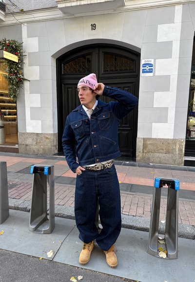 Chaqueta de mezclilla azul oscuro y jeans baggy a juego, con un gorro rosa y botas color beige. Presenta un cinturón de estampado de leopardo. Fondo urbano.