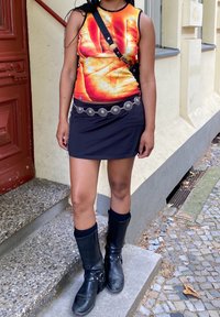 Top de tanque laranja brilhante com um gráfico de mão, combinado com uma mini saia preta. Finalizado com botas pretas até o joelho e um cinto com detalhes em prata.