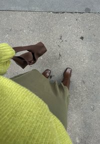 Maglione verde a maglia, vestito verde oliva, stivali in pelle marrone con punta quadrata e una borsa marrone, tutti posizionati su un pavimento texture.