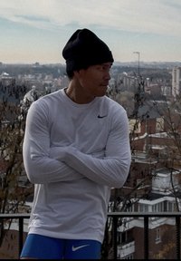 Homme en chemise blanche à manches longues et short bleu, les bras croisés, portant un bonnet noir, debout à l'extérieur sur un balcon surplombant le paysage urbain.