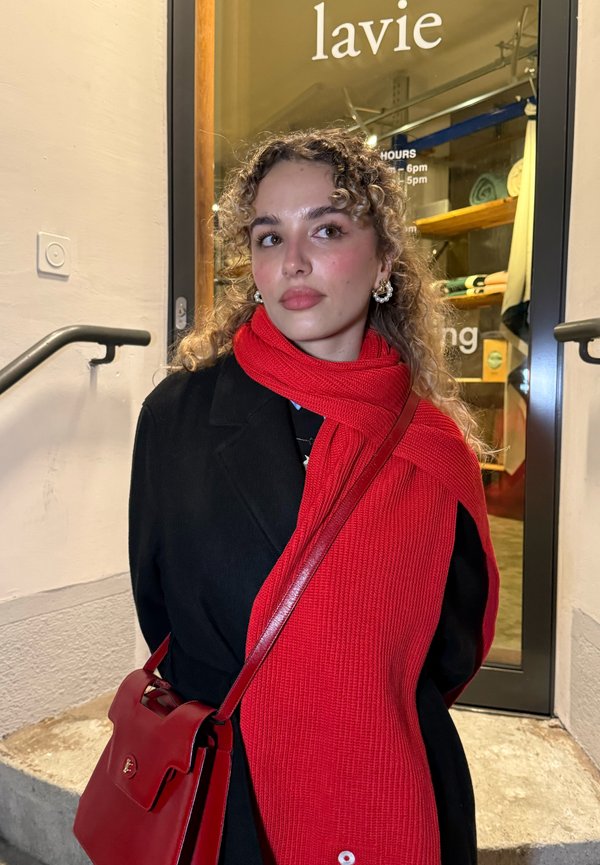 Jeune femme aux cheveux bouclés portant une écharpe rouge, un manteau noir et un sac rouge, debout près d'une porte vitrée avec des étagères à l'intérieur d'un magasin.