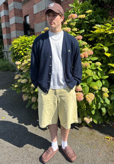Cárdigan azul marino sobre una camiseta blanca, pantalones cortos amarillos, zapatos marrones sin cordones y una gorra marrón. Rodeado de follaje verde y flores rosas.