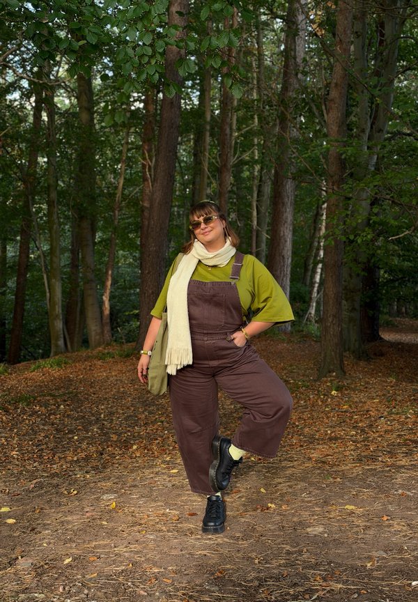 Una mujer con gafas de sol, camisa verde, peto marrón y bufanda color crema posa con una pierna levantada en un bosque frondoso.