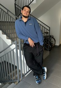 Világoskék polárzippes pulóver, fekete cargo nadrág és színes sportcipők. Lépcsőn áll a szürke korlát mellett, a háttérben kerékpárok.