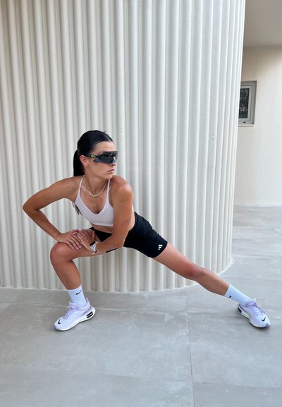 Conjunto atlético que incluye un sujetador deportivo blanco, pantalones cortos negros con un logotipo y calcetines blancos. Los accesorios incluyen gafas de sol y un collar de cadena.