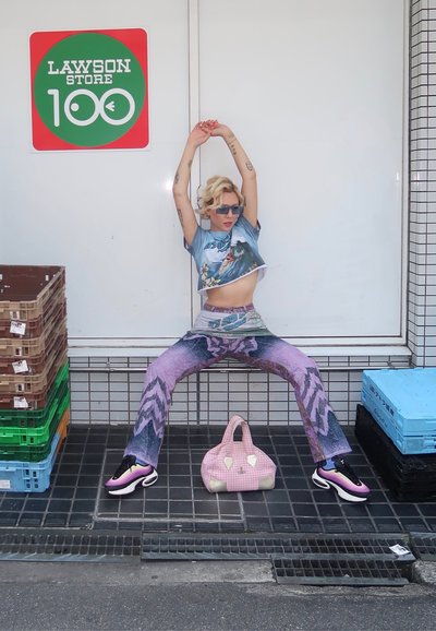 Persona vestida con una camiseta gráfica azul de corte corto, jeans de campana multicolores, zapatillas negras y rosas, sosteniendo un bolso de mano de patrón rosa, posando al aire libre.