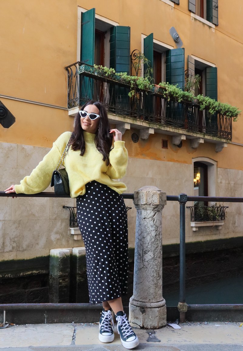 Donna con occhiali da sole bianchi, maglione giallo, gonna nera a pois e scarpe da ginnastica che si appoggia a una ringhiera vicino a un canale, con un edificio giallo e persiane verdi sullo sfondo.
