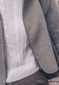 Grå ytterjakke med glidelås over en hvit kabelstrikket genser med ribbet kant, som viser jevn tekstur og kontrasterende farger.