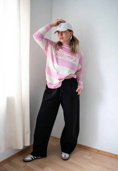 Camisa de manga larga a rayas en rosa y beige con el texto "CALI SPORTS", combinada con pantalones holgados negros y zapatillas grises. Se lleva una gorra blanca de béisbol.