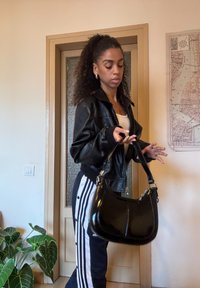 Schwarze Handtasche aus Lackleder mit geschwungener Form und glänzendem Finish, ausgestattet mit silbernen Beschlägen und einem einzelnen Schulterriemen.