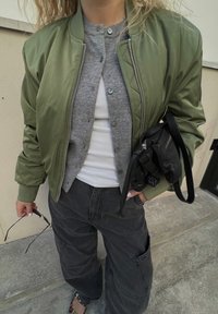 Olive green bomber jacket, grey cardigan, white tank top, dark cargo pants, black bag with straps, sunglasses in hand, textured materials.