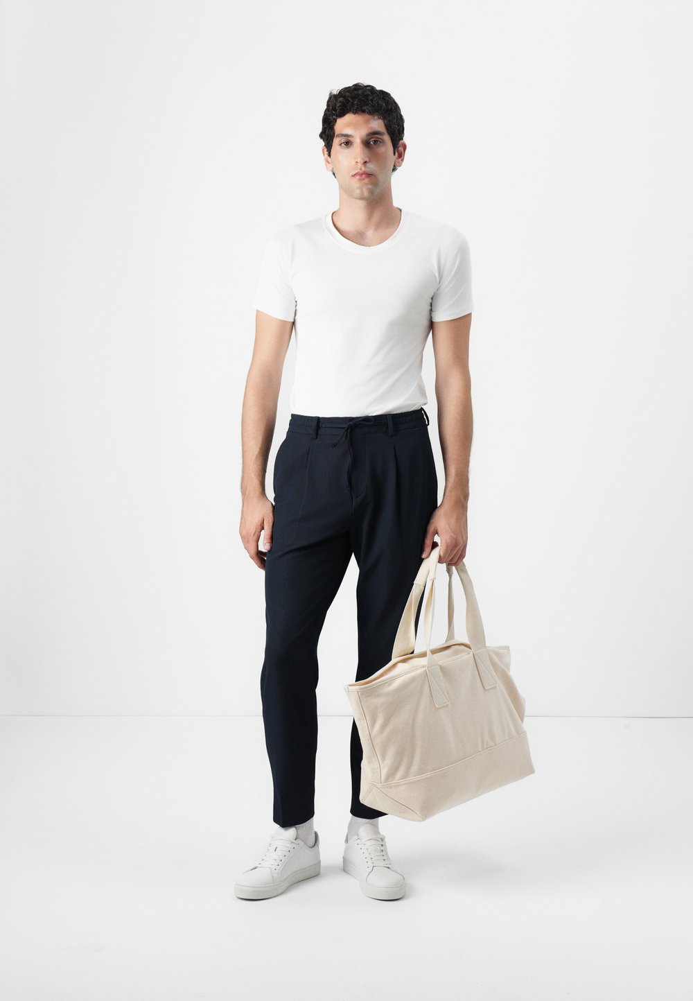 T-shirt blanc en coton, pantalon marine et baskets blanches. Tient un sac en toile beige avec deux poignées robustes. Look simple et moderne.