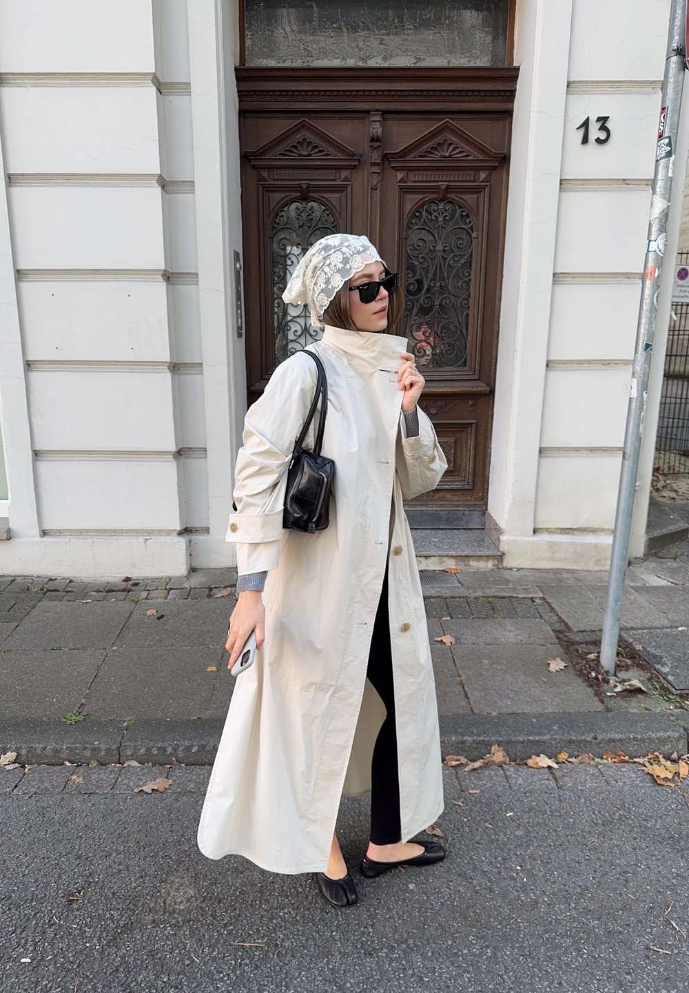 Beige trenčkot, lehký materiál, detaily na knoflících. Černá kabelka, ploché černé boty, šedý dlouhý rukáv, vzorovaný šátek na hlavu, tmavé sluneční brýle.