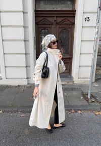 Beige trenčkot, lehký materiál, detaily na knoflících. Černá kabelka, ploché černé boty, šedý dlouhý rukáv, vzorovaný šátek na hlavu, tmavé sluneční brýle.