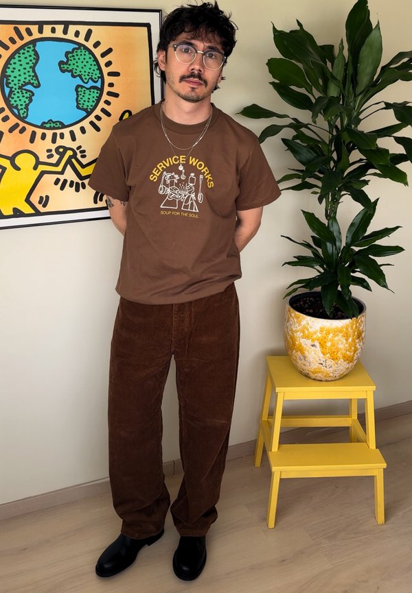 Un homme portant des lunettes et des vêtements bruns se tient à côté d’une grande plante en pot et d’une illustration colorée encadrée.