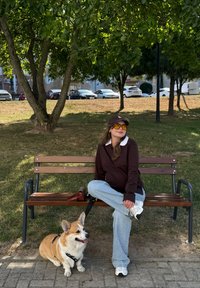 Ung kvinde i afslappet tøj og gule solbriller sidder med krydsede ben på en bænk i parken, med en corgi-hund i snor ved siden af hende.