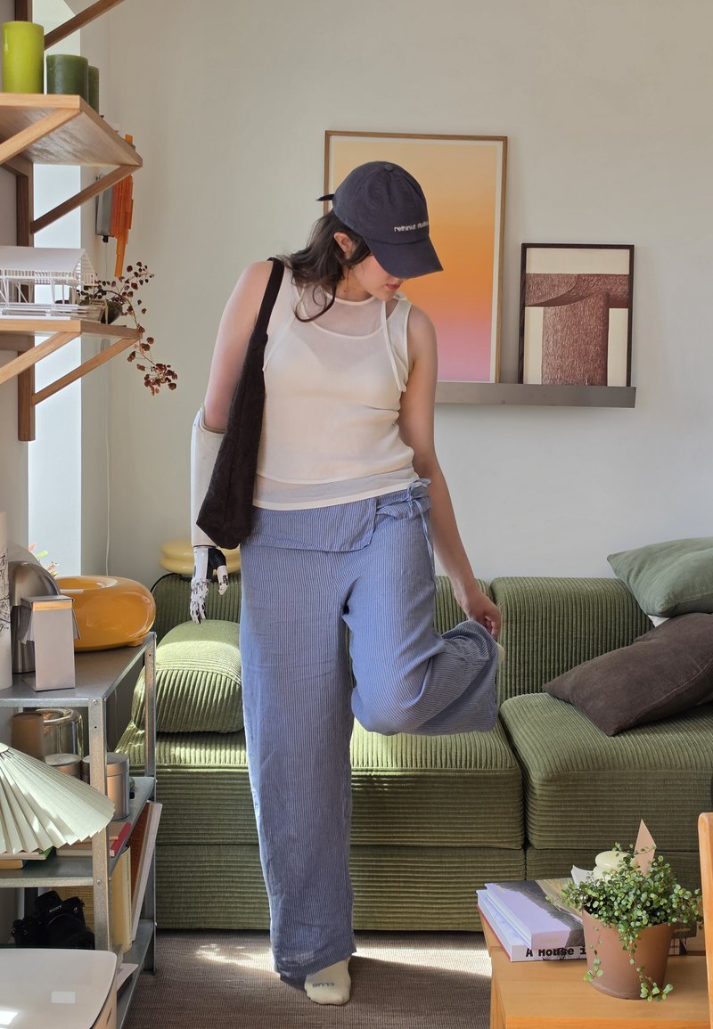 Personne avec un bras prothétique portant une casquette, un haut blanc sans manches et un pantalon bleu se tient dans un salon avec un canapé vert et des œuvres d'art encadrées.