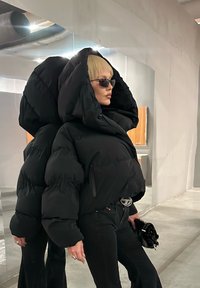 Pessoa com cabelo loiro curto, vestindo um grande casaco acolchoado preto, óculos de sol e calças pretas, em pé ao lado de uma parede refletiva no interior.