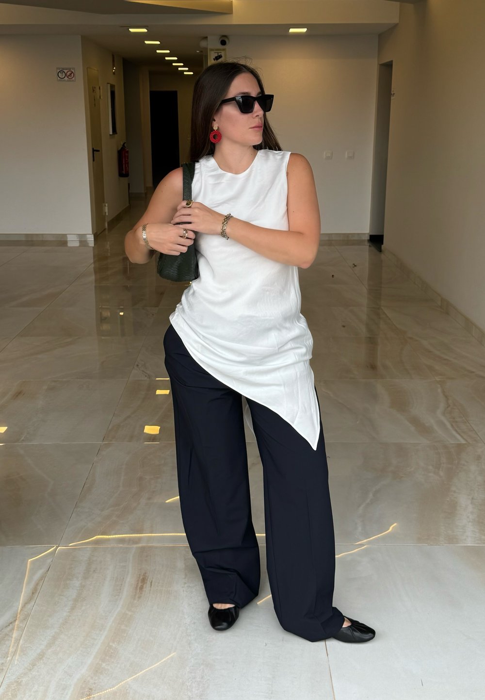 Hvit ermeløs topp med asymmetrisk kant, parret med løse marineblå bukser. Svarte leiligheter og en grønn håndveske. Røde sirkulære øredobber som detalj.