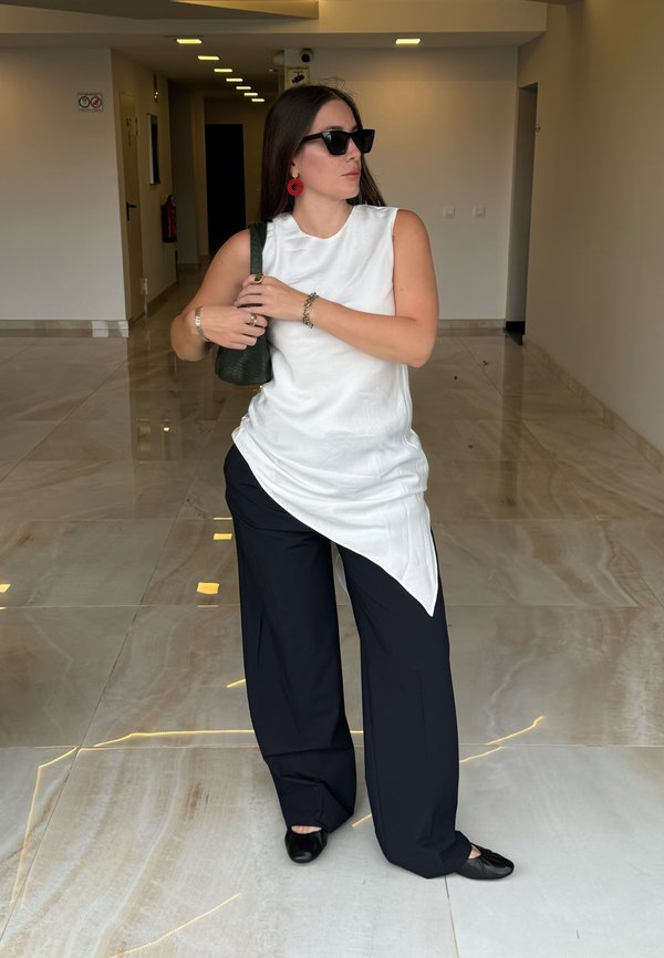 Weißes ärmelloses Top mit asymmetrischem Saum, kombiniert mit lockeren, marineblauen Hosen. Schwarze Ballerinas und eine grüne Handtasche. Rote, runde Ohrringe als Detail.