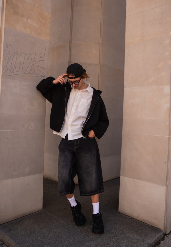 Persona joven con gorra negra, gafas, chaqueta negra, camisa blanca y pantalones cortos amplios, apoyada contra una pared de piedra beige con grafitis.