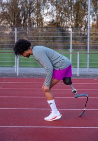 Atletisk mann iført en grå langermet skjorte og lilla shorts, med et svart protetisk ben, på en rød løpebane, forbereder seg på å løpe.