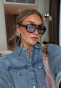 Sunglasses in oversized tartaruga, camicia di jeans con dettagli in bottone argentato e una sciarpa rosa a fantasia con frange. Capelli sciolti.