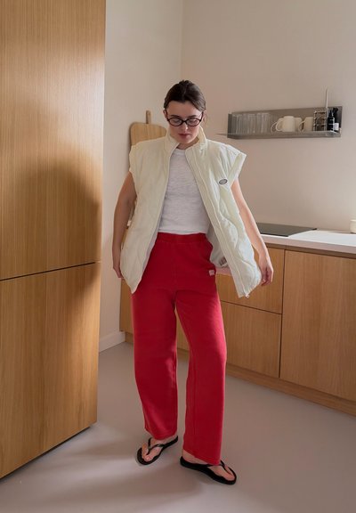 Persona con gafas, chaleco acolchado blanco sin mangas, camisa blanca, pantalones rojos y sandalias negras de goma de dedo de pie en una cocina moderna.