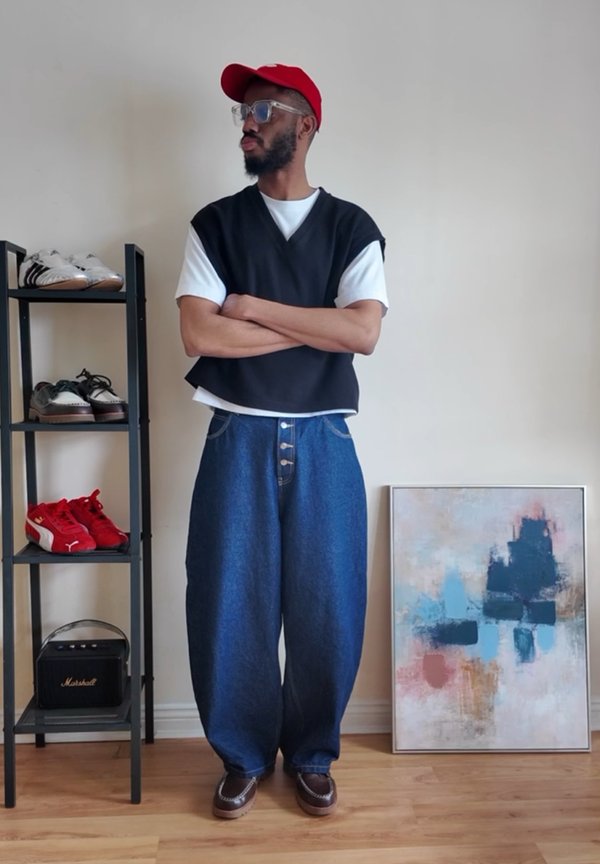 Man wearing red cap, glasses, black sleeveless top over white t-shirt, wide blue jeans, standing with arms crossed next to shoe rack and abstract painting.