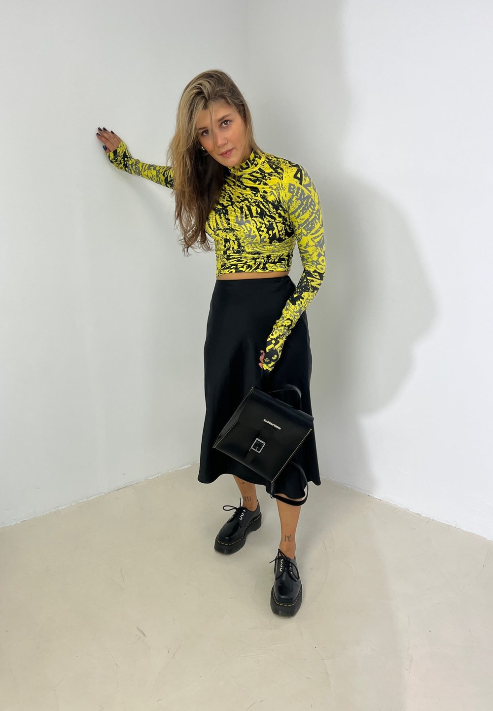Long-sleeved cropped top in yellow featuring black abstract patterns, teamed with a black satin skirt, black platform shoes, and a black handbag.