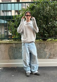 Pull en maille grise avec col polo, jean oversize bleu clair, et chaussures grises sans lacets. Cheveux coiffés en boucles lâches, debout en extérieur devant un fond de verdure.